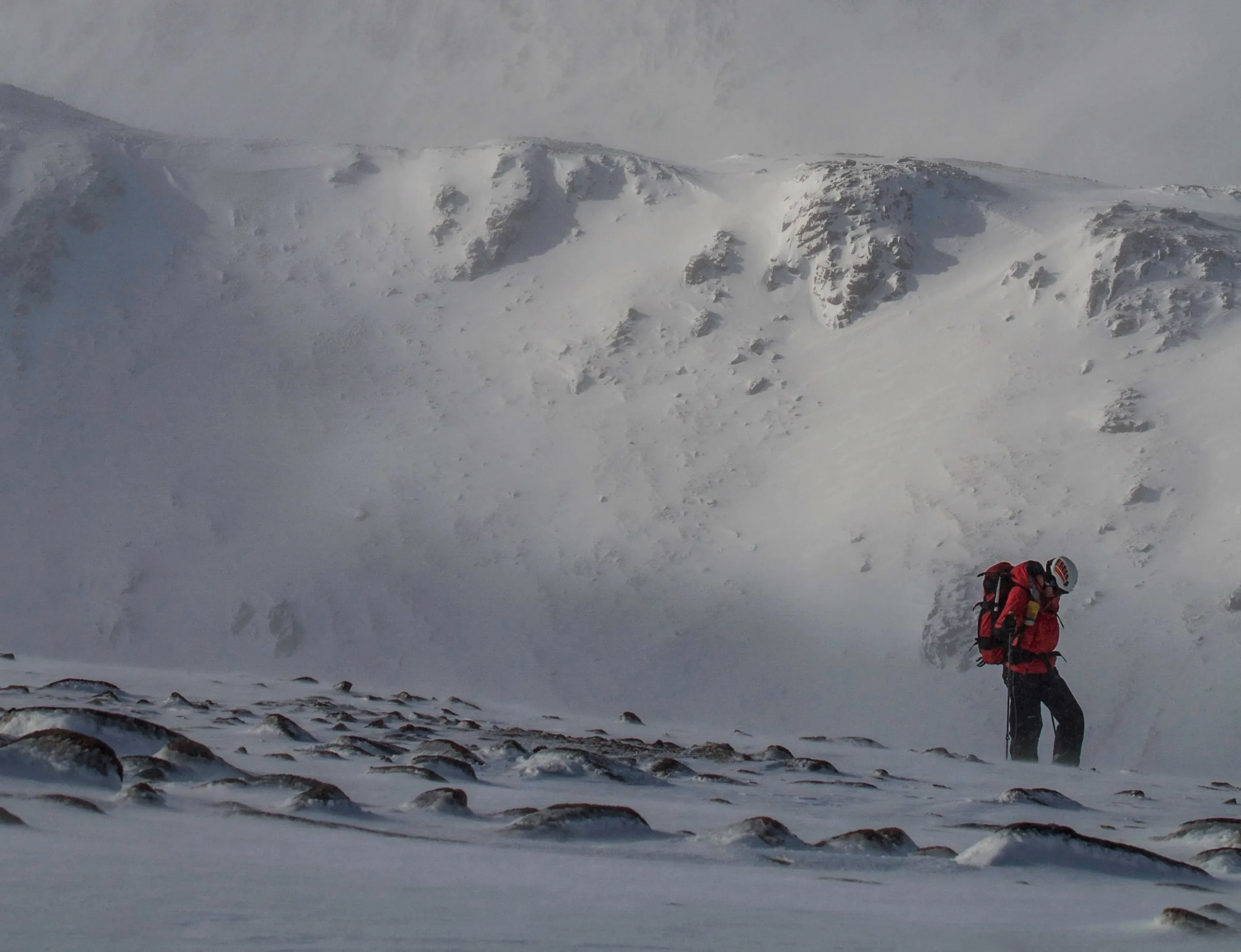 A lone mountain rescue team member crossing snow below a steep ridge near the Devil's Point
